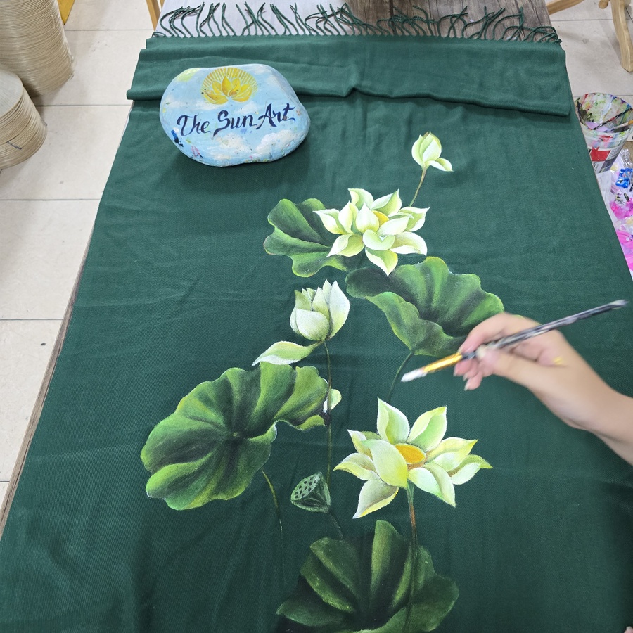 Artist hand-painting white lotus flowers on a dark green scarf at The Sun Art studio
